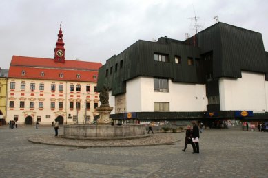 Věda, architekti a památky - <B>Regenerace a dialog počesku</B>: Zdeněk Sklepek, obchodní dům Prior, Jihlava, 1978-83, foto 2008. Nositel titulu CSc. rozčlenil hmoty pomocí „domovních“ oken a falešných střech tak dětinsky, až se pozorovateli téměř nechce věřit, že za tím nestojí nějaký obzvlášť rafinovaný koncept. Možná první příklad tzv. podnikatelského baroka u nás. Stromy zbytečně zakrývají historické fasády, zatímco obchodní dům a zásobovací rampy rozrývající povrch náměstí zůstávají odkryté - foto: Martin Horáček